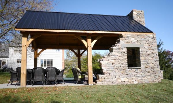 12 x 20 Custom Timberframe Pavilion with Stone wall A 12 x 20 timber frame pavilion with a black metal roof, stone fireplace, and a patio set with six chairs is shown on a grassy lawn. A house is visible in the background.