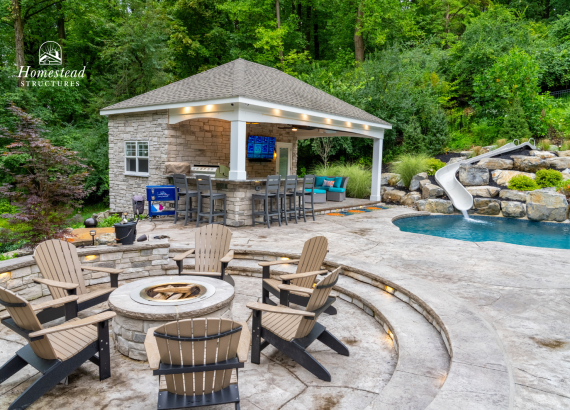 Stunning backyard with custom-built luxury pool house, outdoor kitchen, inviting swimming pool with waterslide, cozy firepit seating area, and lush greenery, ideal for entertaining and relaxing.