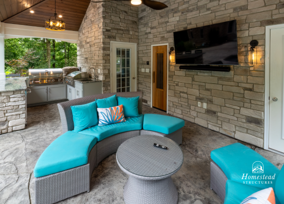 Interior of a luxurious pool house featuring elegant stone walls, comfortable turquoise seating, mounted flat-screen TV, sleek ceiling fan, and an adjacent outdoor kitchen with stainless steel appliances.