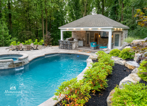 Elegant stone pool pavilion with spacious entertainment area including an outdoor kitchen, built-in grill, comfortable seating, fire pit area, and integrated landscaping, perfect for gatherings