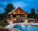 A backyard features a 16' x 14'  timber frame pavilion with a lit fireplace, surrounded by seating, next to a curved swimming pool at dusk. Trees and a house are in the background.